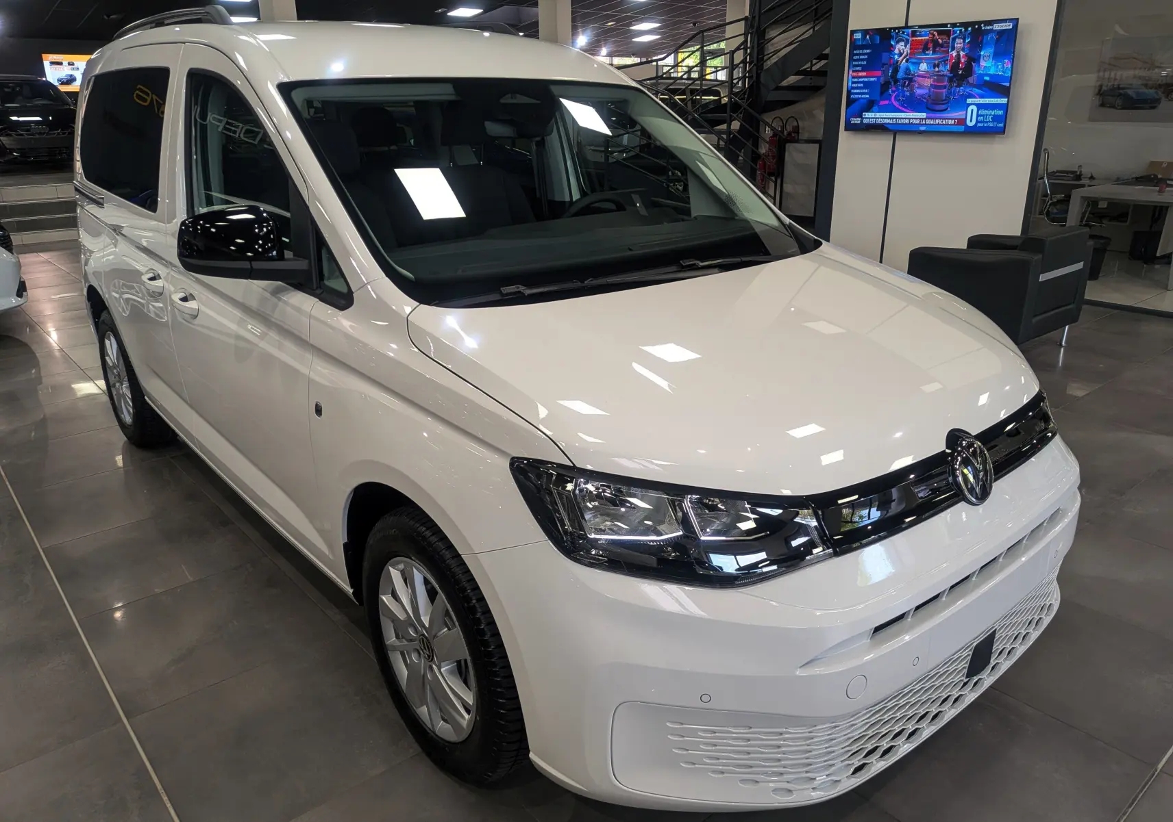 Vue 3/4 avant d'un Volkswagen Caddy blanc 2025 en showroom, avec calandre noire et jantes argentées.