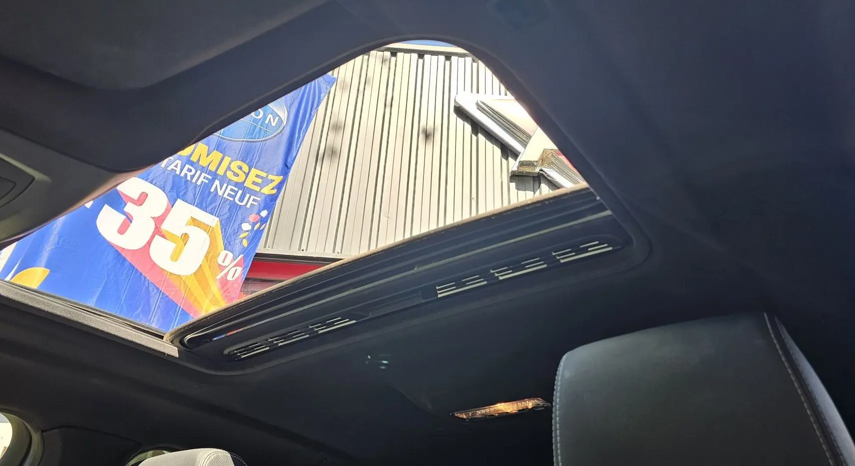 Toit ouvrant panoramique ouvert vu de l'intérieur d'une BMW X4 xDrive20dA noire, avec ciel et bannière publicitaire en arrière-plan.