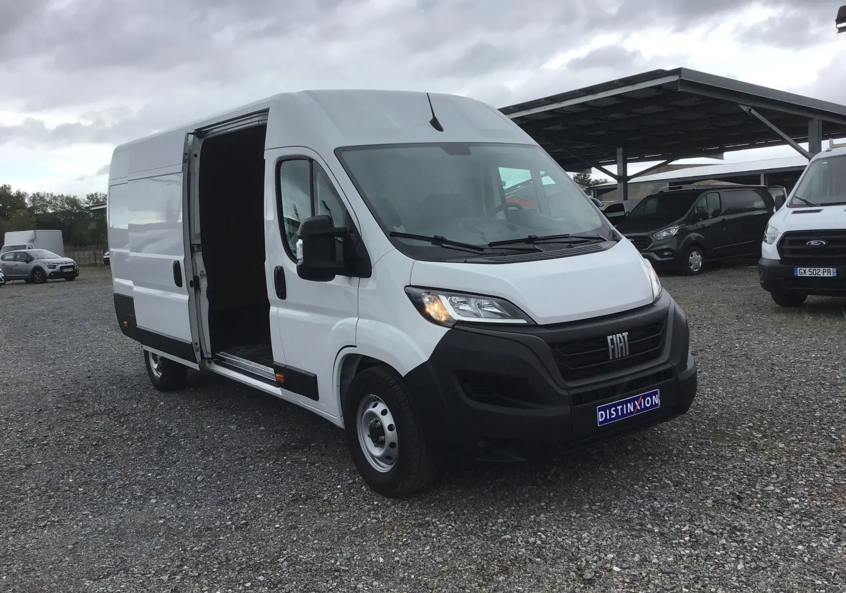Vue 3/4 avant gauche du Fiat Ducato Fourgon blanc 2024 avec porte latérale ouverte sur parking gravillonné.