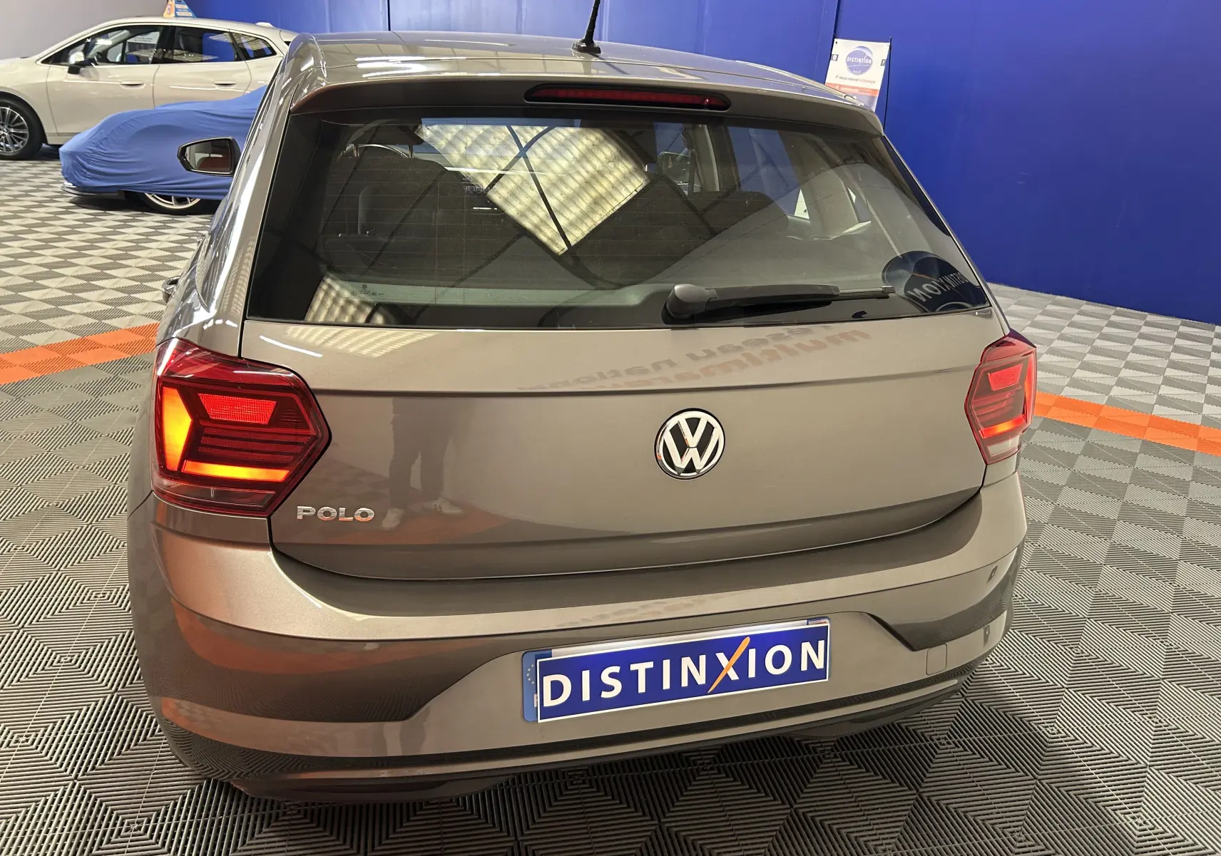 Vue arrière d'une Volkswagen Polo gris métallisé avec feux allumés, stationnée en intérieur sur sol damier.