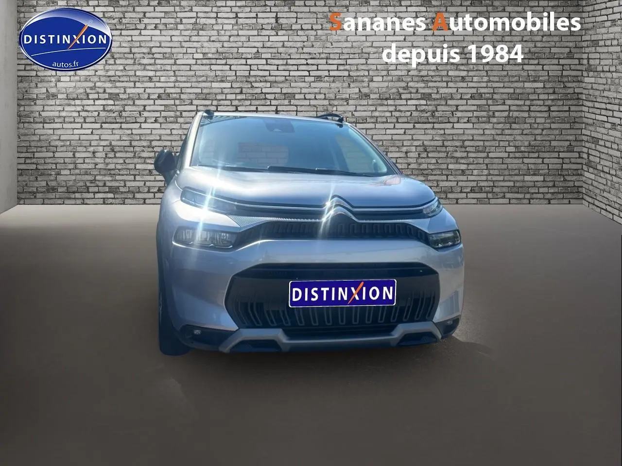 Vue frontale d'un Citroën C3 Aircross gris foncé avec projecteurs allumés, sur fond de mur en briques.