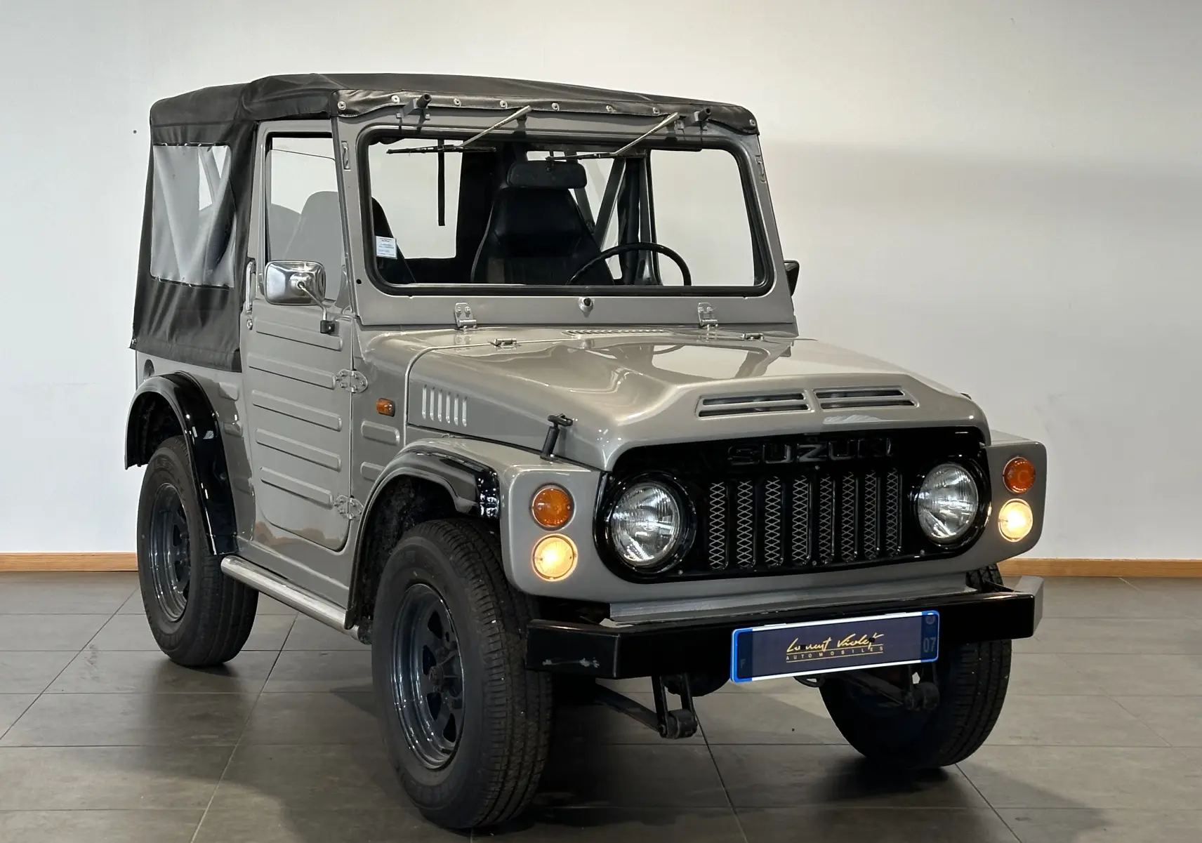 Suzuki Across LJ80 Cabriolet gris vu en 3/4 avant droit avec toit souple noir et phares allumés.