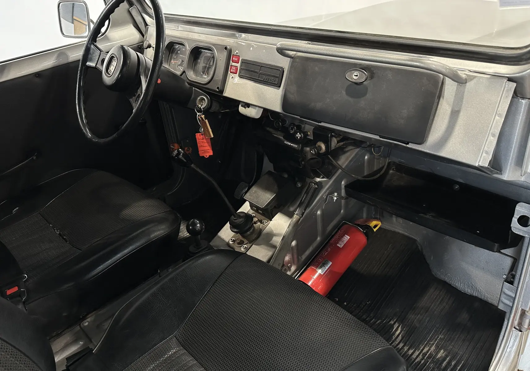 Intérieur du Suzuki Across LJ80 Cabriolet gris 1981 vu côté conducteur, avec tableau de bord simple et extincteur au sol.