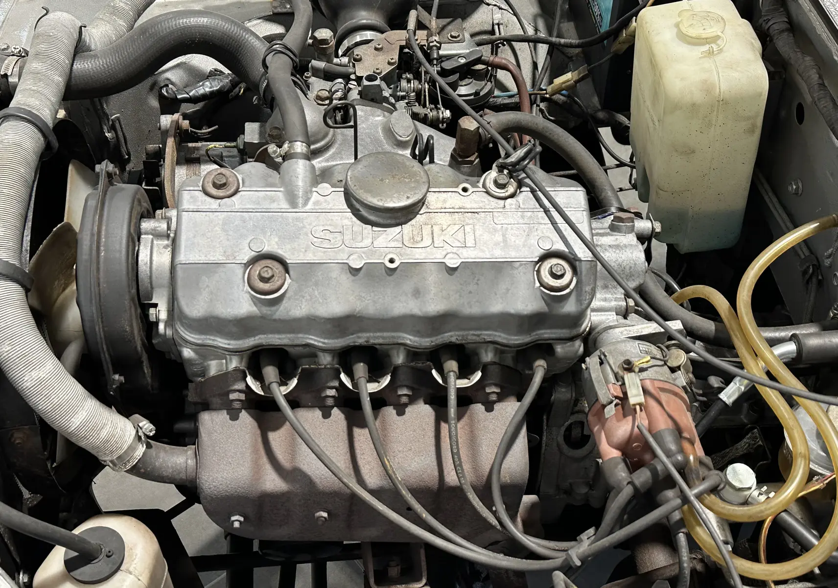 Gros plan sur le moteur essence gris marqué SUZUKI du Suzuki ACROSS LJ80 Cabriolet 1981, avec câbles et durites visibles.