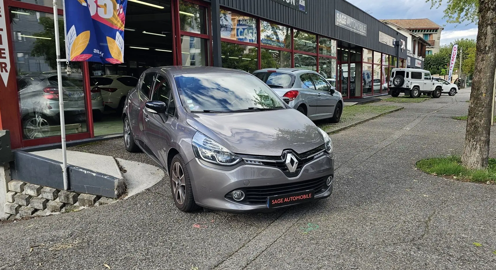 Renault Clio IV grise vue 3/4 avant droit, garée devant un garage avec plaque SAGE AUTOMOBILE.
