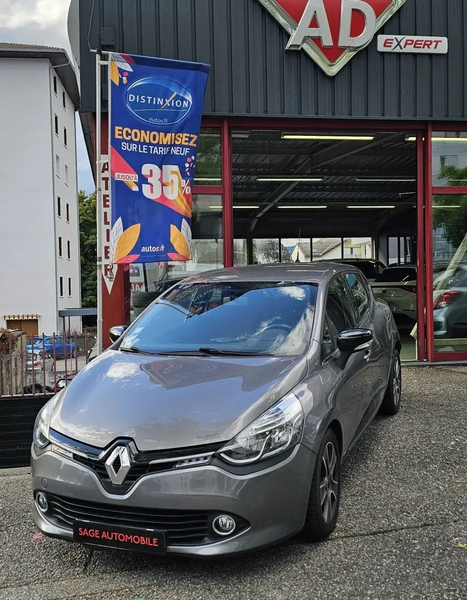 Renault Clio IV grise vue 3/4 avant droit devant un garage AD Expert avec drapeau promotionnel bleu.