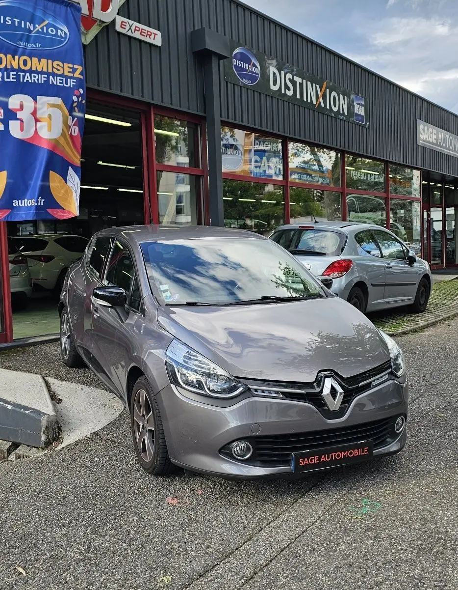 Renault Clio IV grise vue 3/4 avant droit devant un garage avec enseignes et panneau publicitaire coloré.