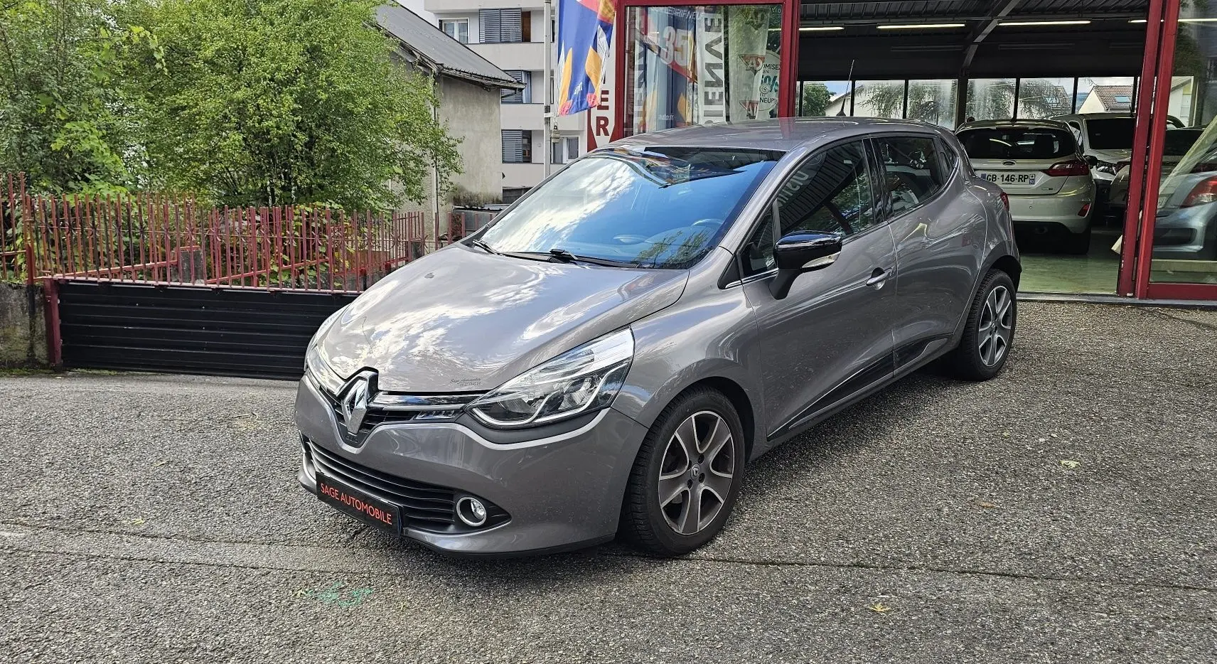 Renault Clio IV grise vue 3/4 avant droit, garée devant un bâtiment avec reflet du ciel sur le capot.