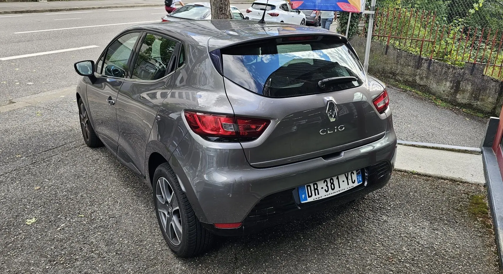 Renault Clio IV grise vue 3/4 arrière côté gauche, avec feux arrière rouges et plaque d'immatriculation française.