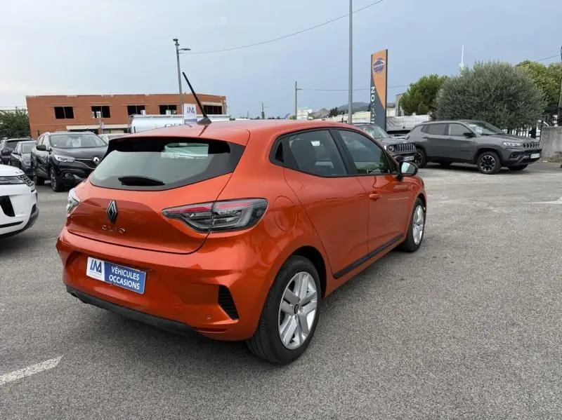 Vue 3/4 arrière droite d'une Renault Clio V orange 2025, version SCE 65 Evolution, garée en extérieur sur parking.