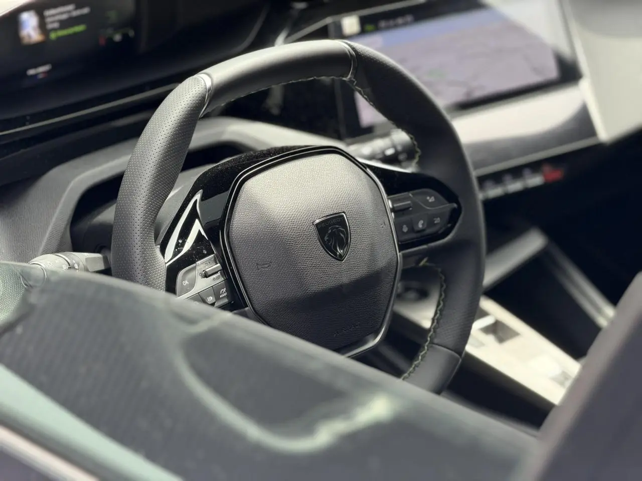 Vue rapprochée du volant compact noir du Peugeot 308 NV Hybrid 145 GT avec surpiqûres vert Adamite et tableau de bord numérique.