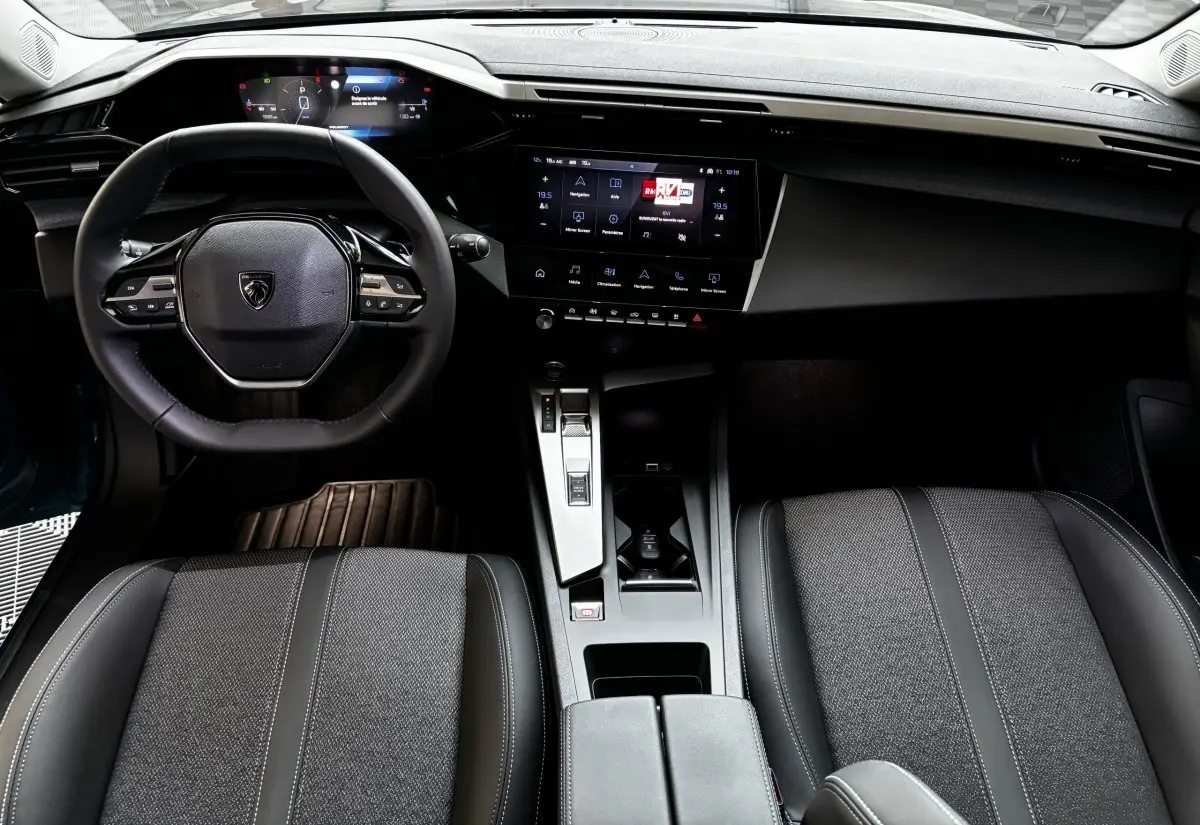 Vue intérieure avant de la Peugeot 308 Hybrid 136 e-DCS6 Allure 2025, volant cuir et écran tactile central noir.