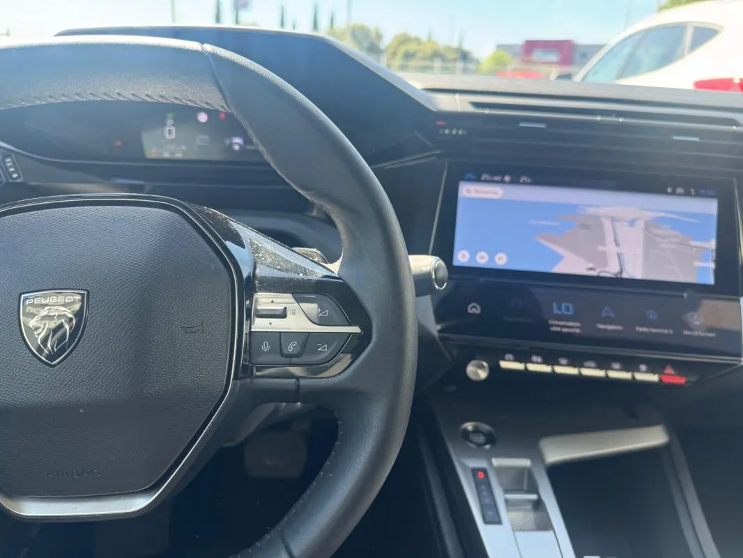 Vue intérieure centrée sur le volant Peugeot gris et le grand écran tactile du tableau de bord avec navigation affichée.