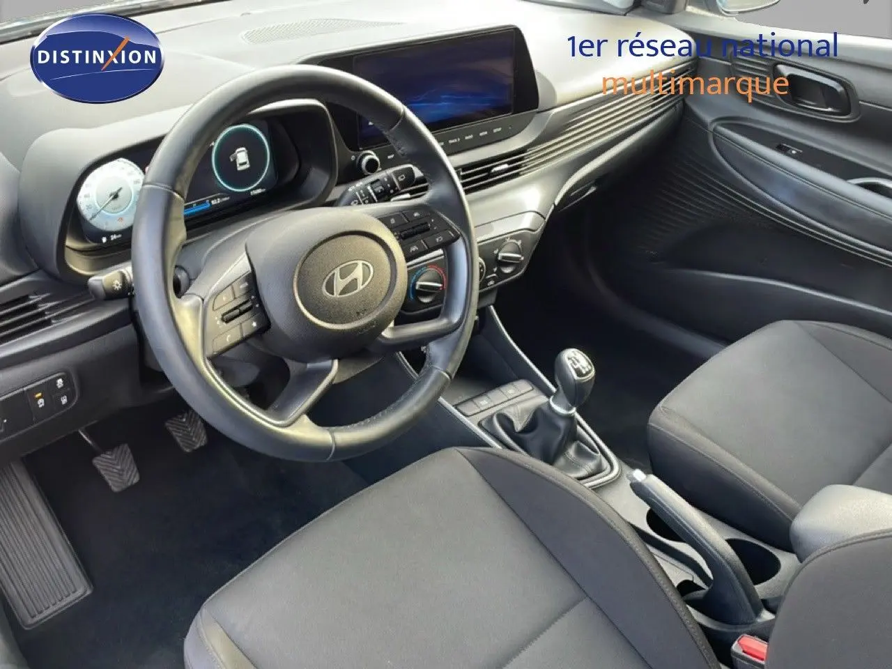 Vue intérieure côté conducteur de la Hyundai i20 2025 avec volant, tableau de bord numérique et levier de vitesses manuel.