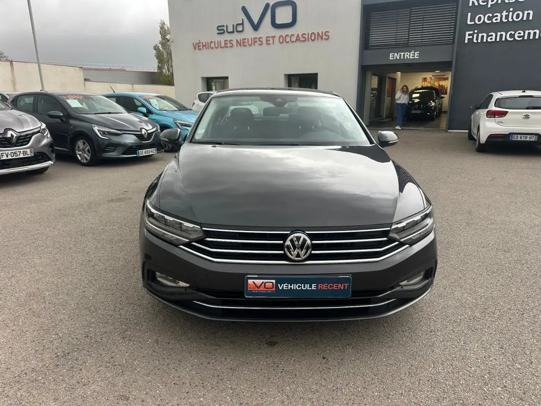 Volkswagen Passat gris foncé vue de face, avec calandre chromée et phares LED allumés sur parking extérieur.