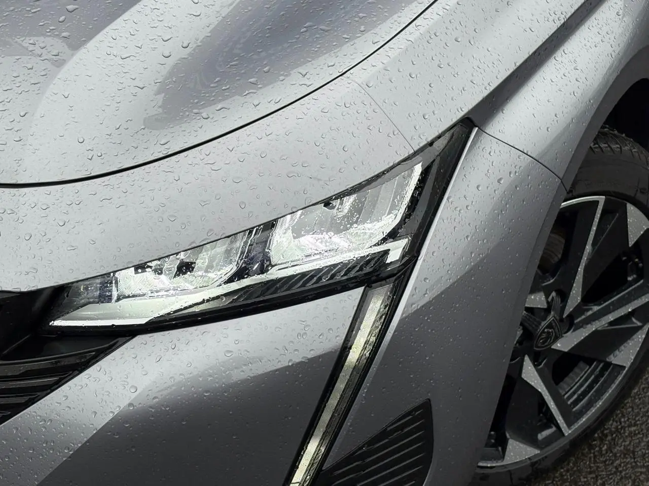 Gros plan sur le phare avant gauche et la roue alliage 17'' d'une Peugeot 308 SW Hybrid gris Artense sous la pluie.