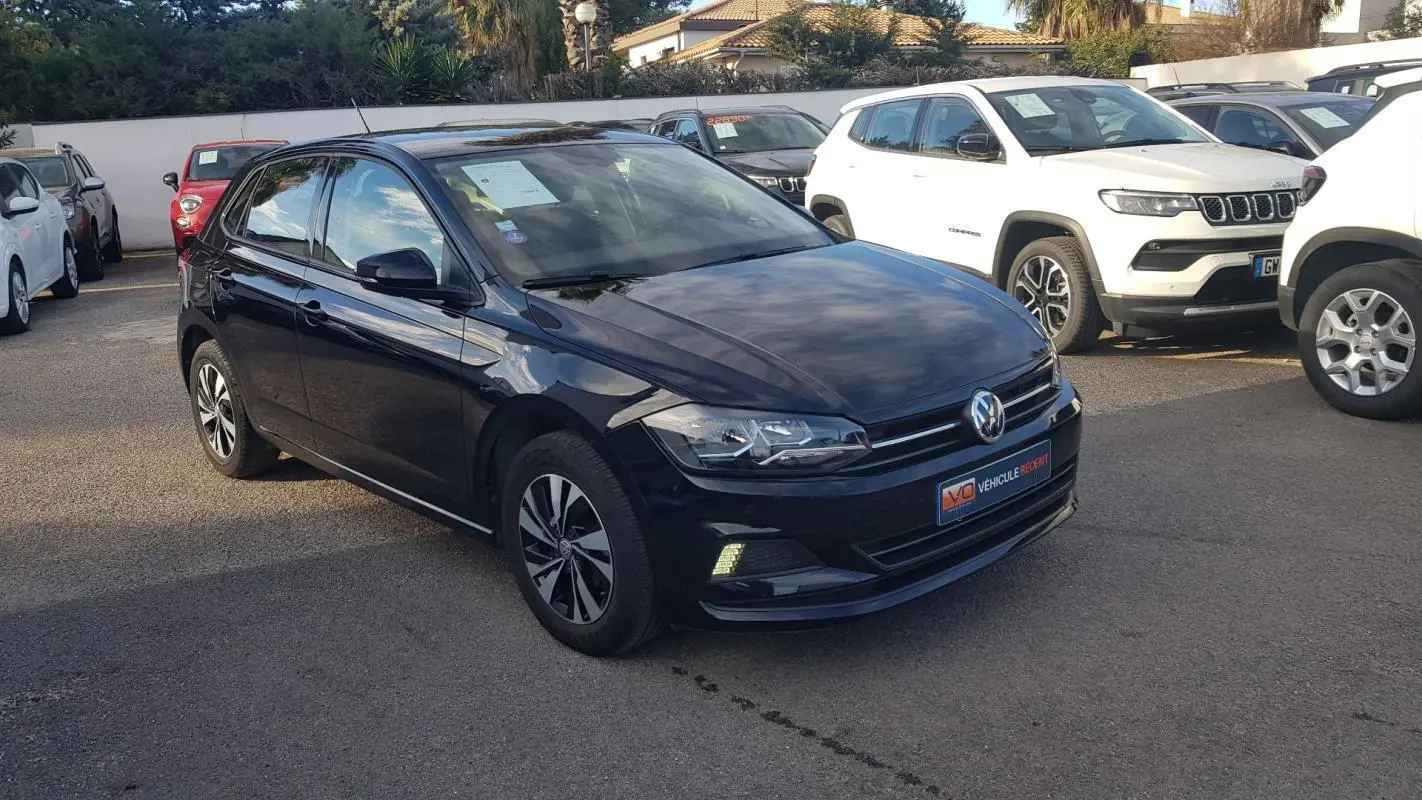 Volkswagen Polo noire vue en 3/4 avant droit avec jantes alliage et feux diurnes allumés sur parking.