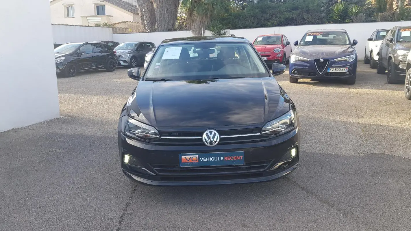 Vue avant d'une Volkswagen Polo noire 2018 avec feux diurnes allumés sur un parking extérieur.