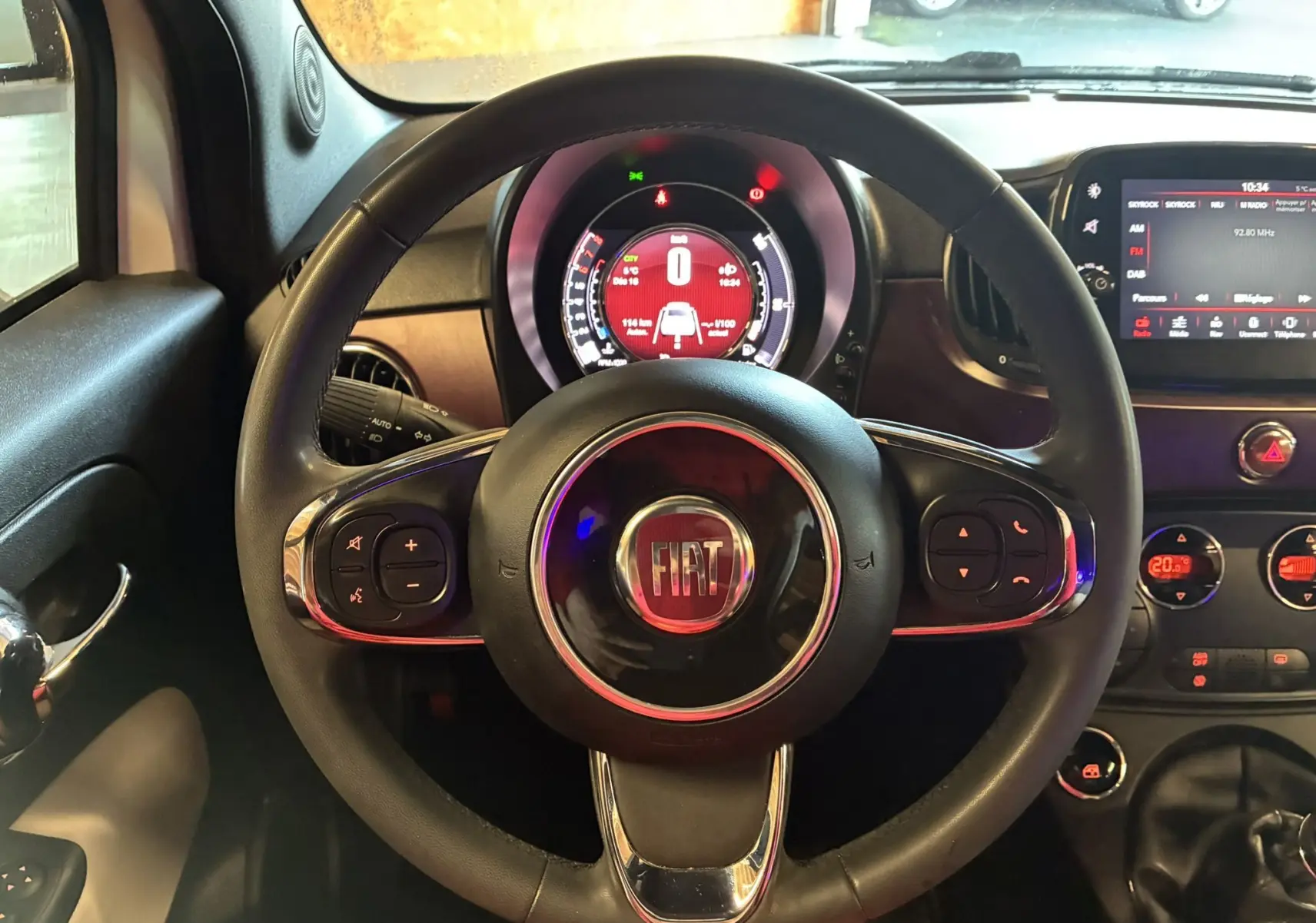 Vue intérieure centrée sur le volant noir de la Fiat 500 blanche, avec tableau de bord numérique et écran tactile visible.