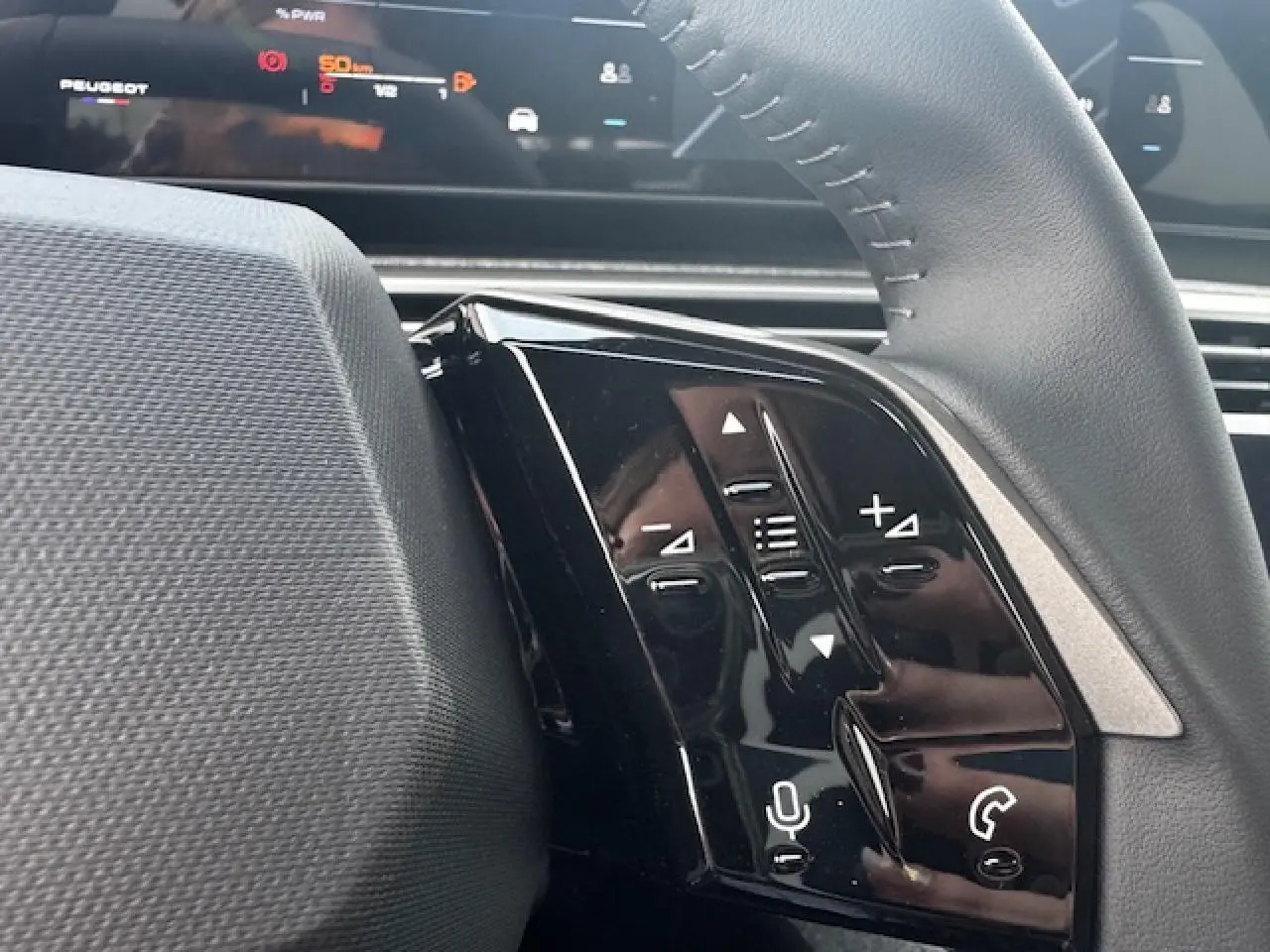 Gros plan sur les commandes gauche du volant cuir noir du Peugeot 5008 bleu Ingaro, avec écran numérique en arrière-plan.