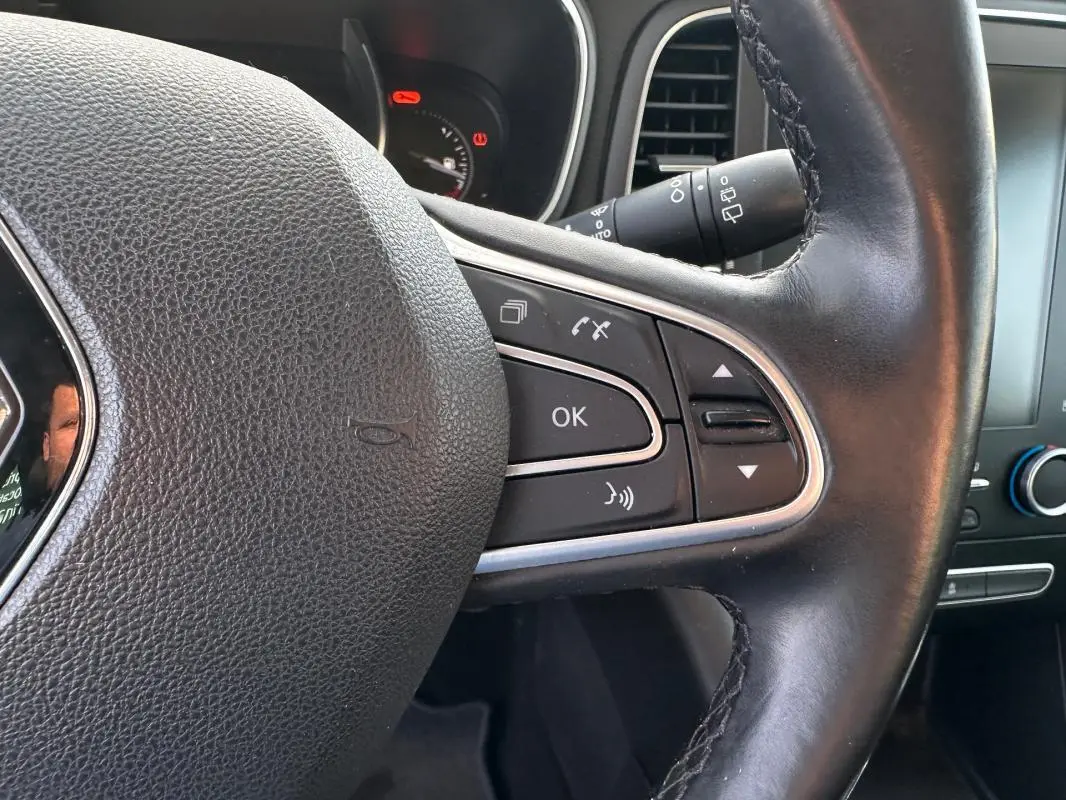 Gros plan sur le volant cuir noir de la Renault Mégane Break gris clair, avec commandes intégrées et tableau de bord partiel visible.