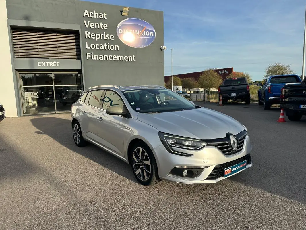 Renault Mégane Break gris clair vue 3/4 avant droit, avec jantes alliage 18 pouces et barres de toit visibles.