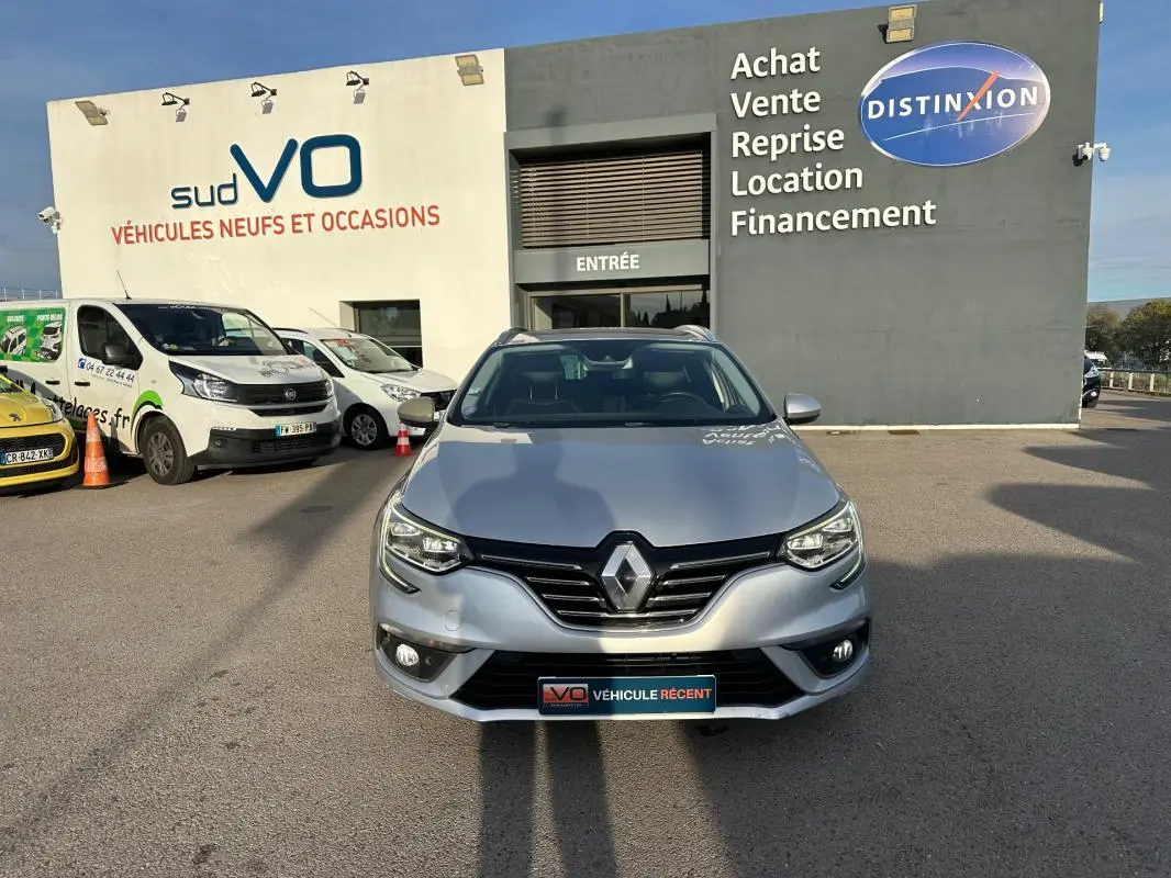 Renault Mégane Break gris clair vue de face, avec barres de toit et phares allumés sur parking de concession.