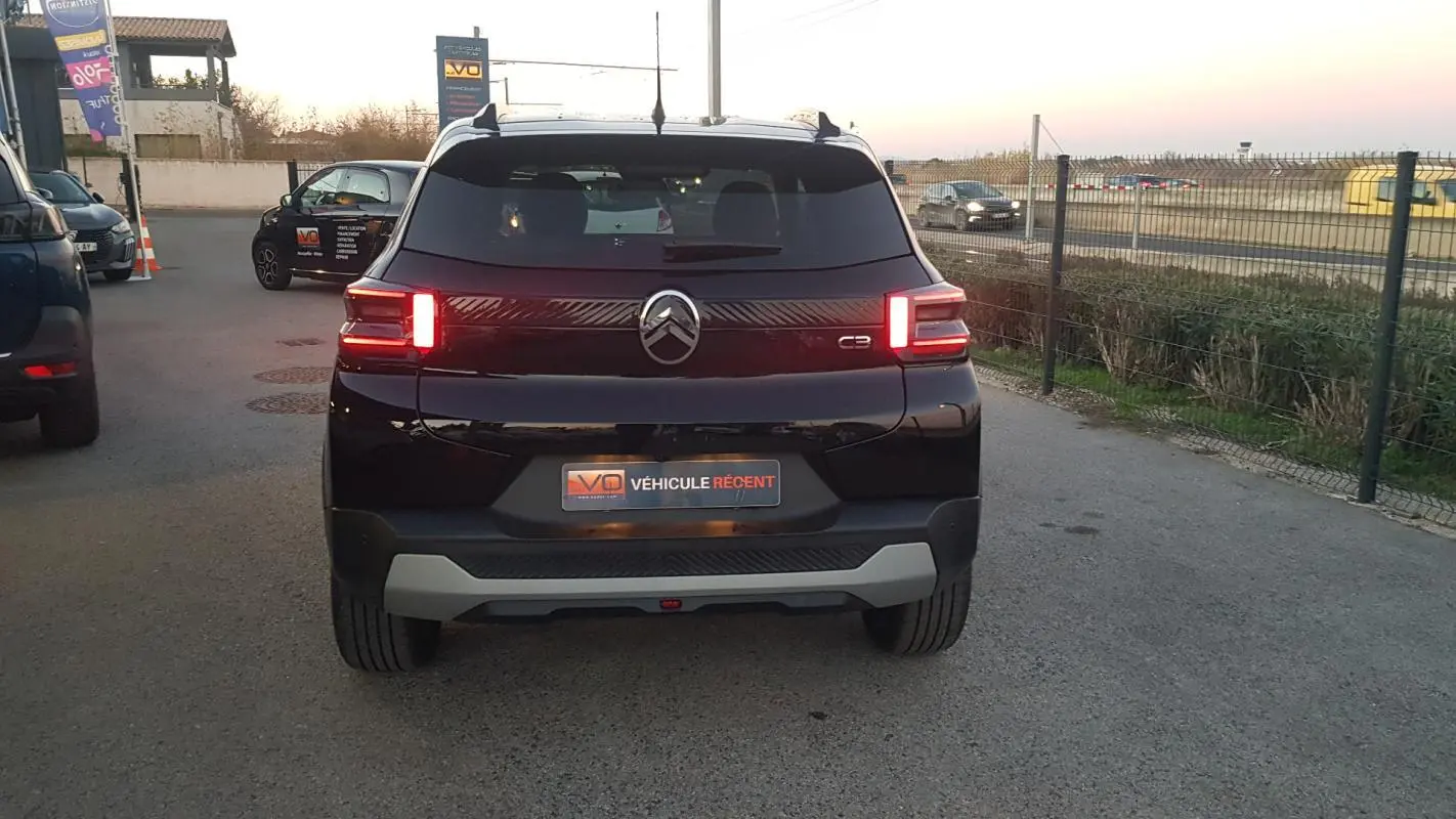 Vue arrière d'une Citroën C3 noire 2025 avec feux allumés et barres de toit visibles en extérieur.