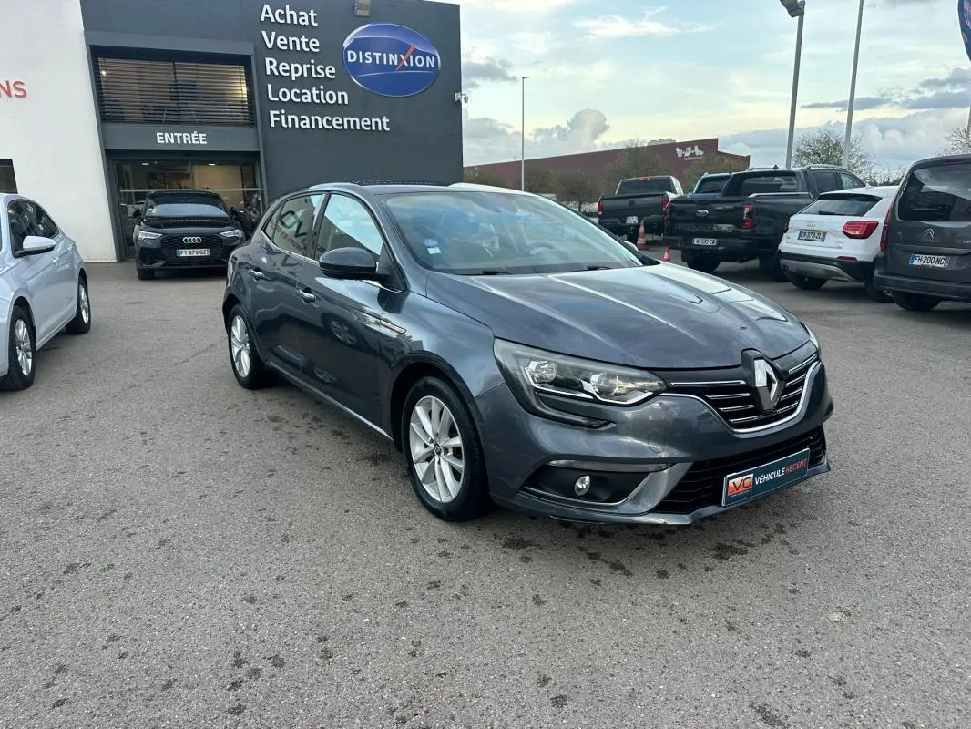 Renault Mégane IV gris foncé vue 3/4 avant avec phares allumés sur parking devant concession.