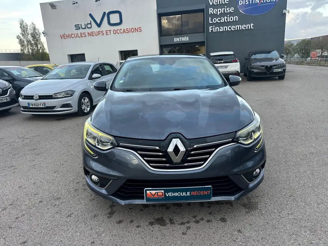 Vue frontale d'une Renault Mégane IV gris foncé avec phares allumés, garée devant un concessionnaire sudVO.