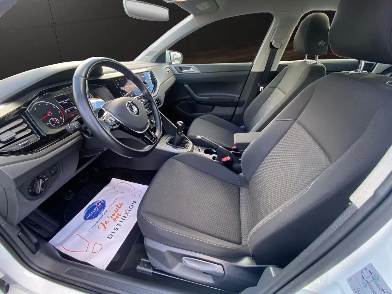 Vue intérieure côté conducteur de la Volkswagen Polo blanche, montrant les sièges noirs et le tableau de bord avec boîte manuelle.