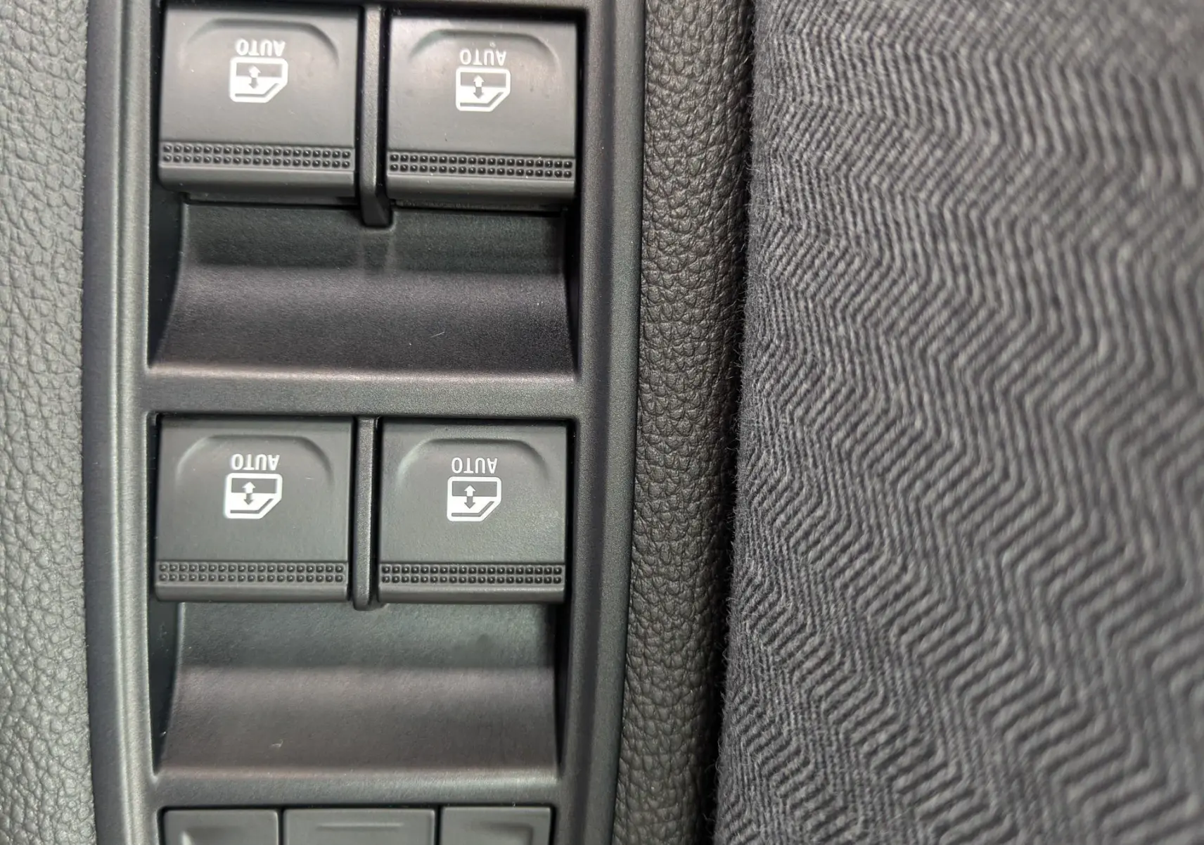 Gros plan sur les commandes de lève-vitres et réglages des rétroviseurs côté conducteur, tissu gris noir visible.
