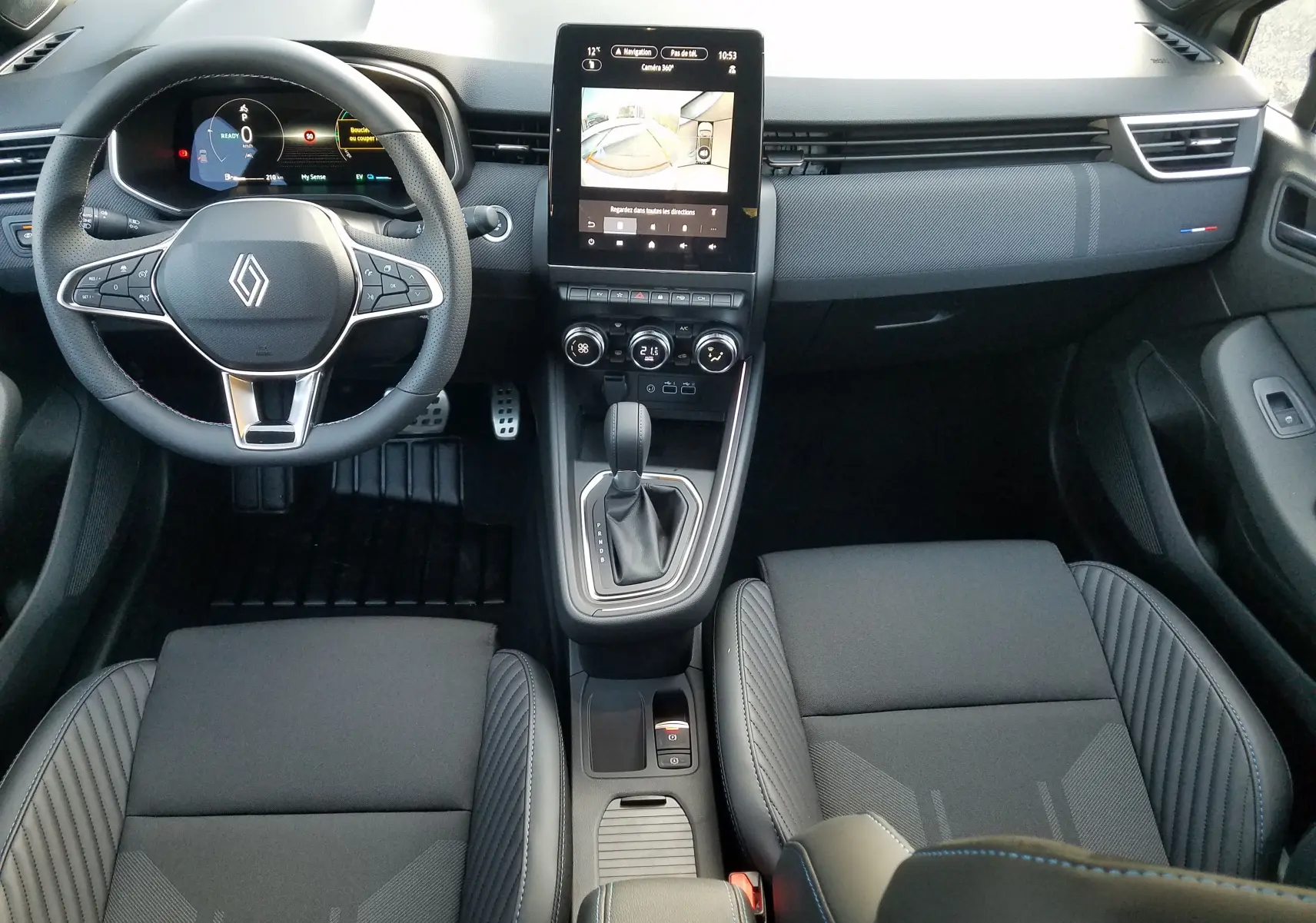 Vue intérieure avant de la Renault Clio V hybride 2025, tableau de bord noir avec écran tactile central et volant multifonction.