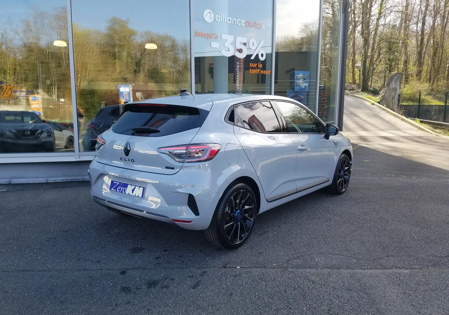 Vue 3/4 arrière droite d'une Renault Clio V gris rafale avec jantes noires et vitres teintées, stationnée devant un showroom.