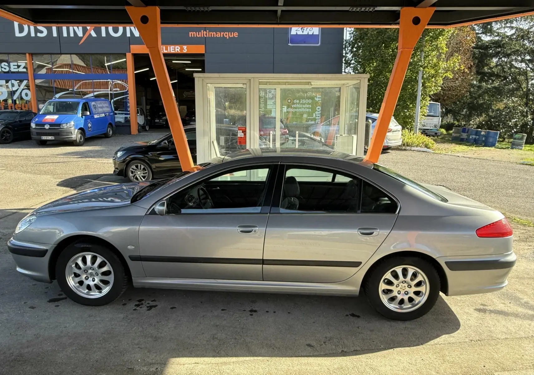 Profil côté gauche d'une Peugeot 607 grise stationnée sous un abri orange avec des jantes alliage visibles.