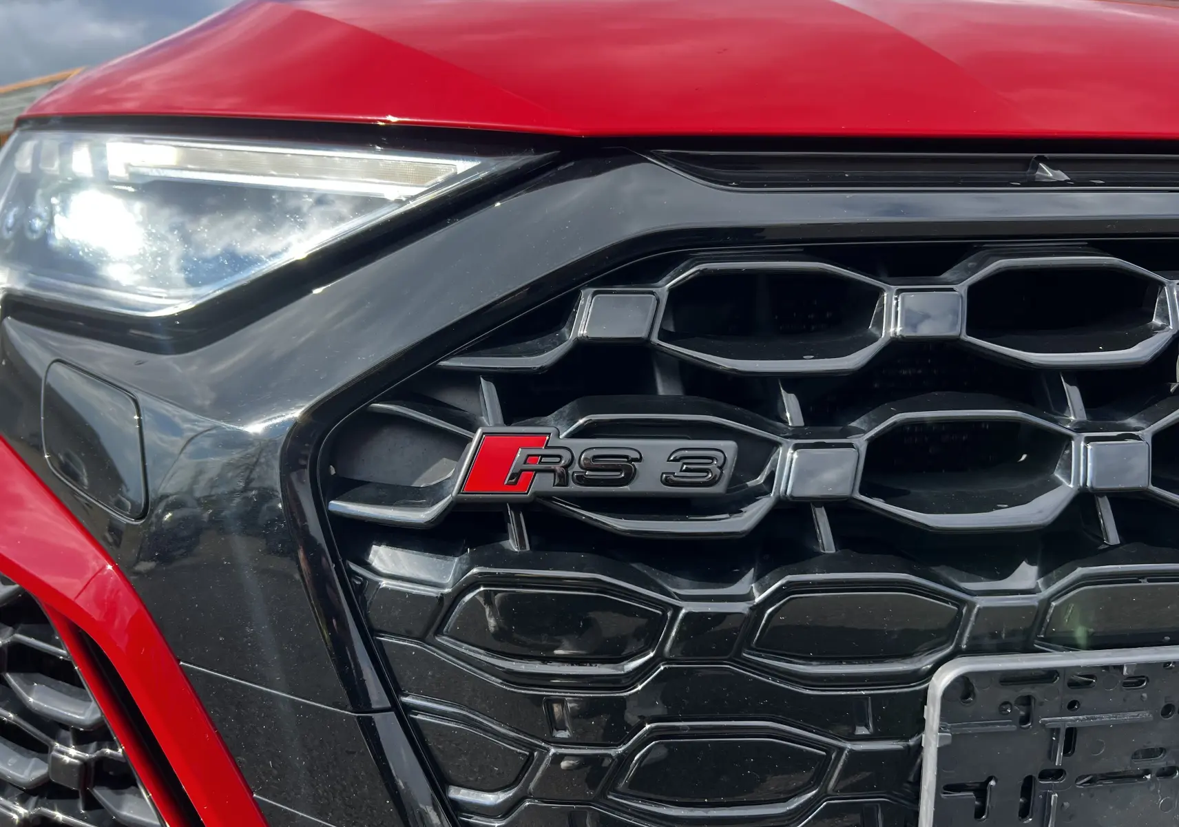 Gros plan sur la calandre noire et le logo RS3 d'une Audi RS3 Sportback rouge, avec un phare avant partiellement visible.