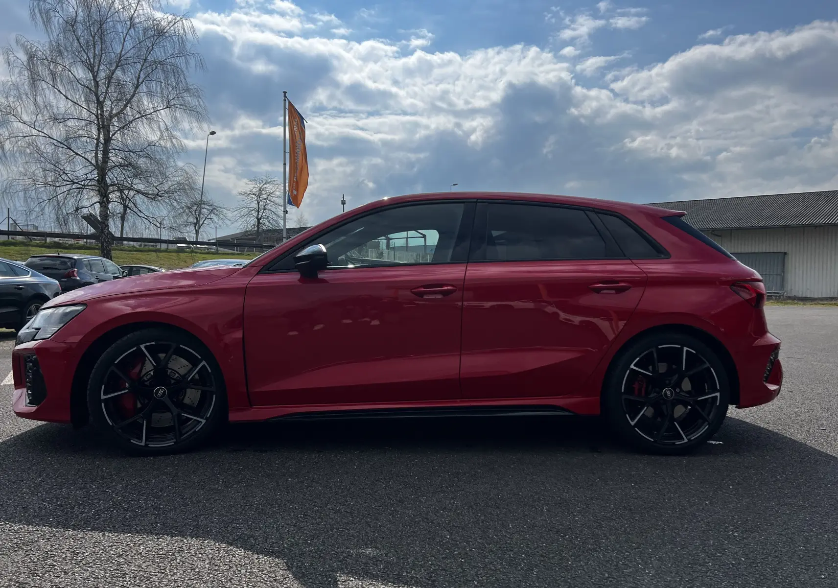 Profil côté gauche d'une Audi RS3 Sportback rouge 2022 avec jantes noires et vitres teintées sur parking extérieur.