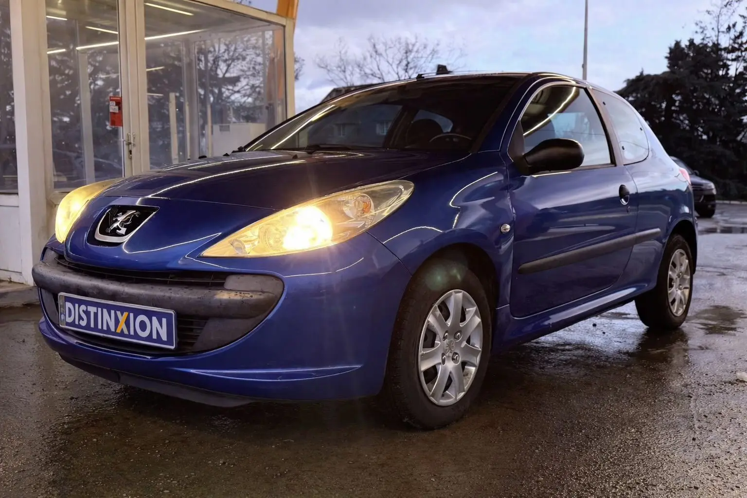 Peugeot 206+ bleu vue 3/4 avant droit avec phares allumés, garée devant un bâtiment vitré.