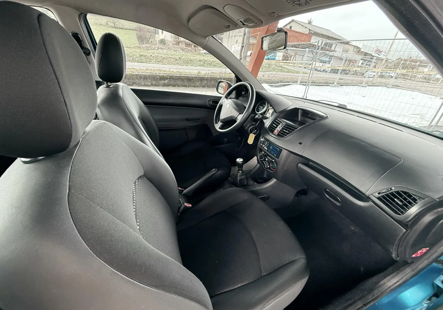 Vue intérieure côté conducteur de la Peugeot 206+ bleue, montrant les sièges noirs en tissu et la console centrale avec boîte manuelle.