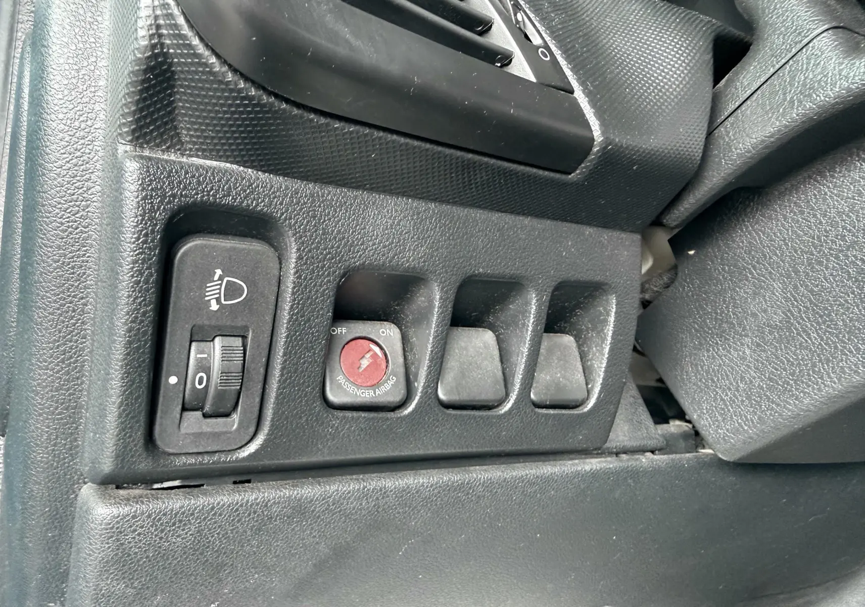 Gros plan sur les commandes noires côté gauche du tableau de bord d'une Peugeot 206+ bleue, avec réglage des phares et airbag passager.