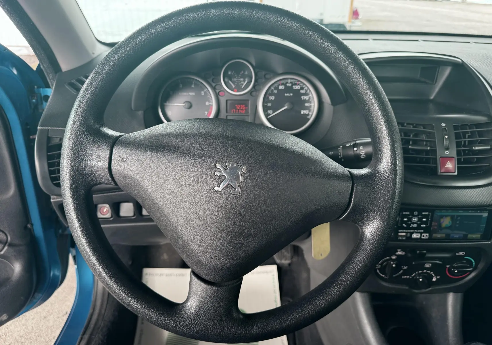 Vue intérieure centrée sur le volant noir de la Peugeot 206+ bleu, avec tableau de bord et console centrale visibles.