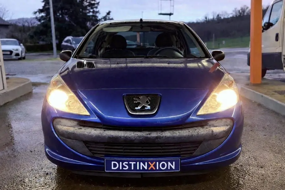 Vue avant d’une Peugeot 206+ bleue de 2011 avec phares allumés et plaque Distinxion visible.