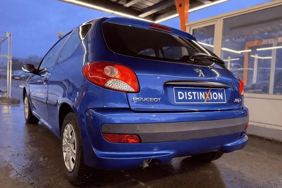 Vue 3/4 arrière gauche d'une Peugeot 206+ bleue garée sous un abri avec plaque Distinxion visible.