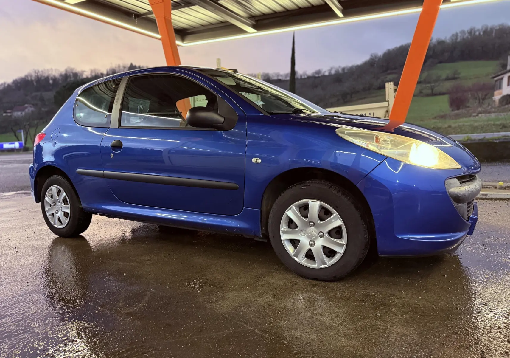 Peugeot 206+ bleu vue de profil côté gauche sous abri, phare avant allumé sur sol mouillé.