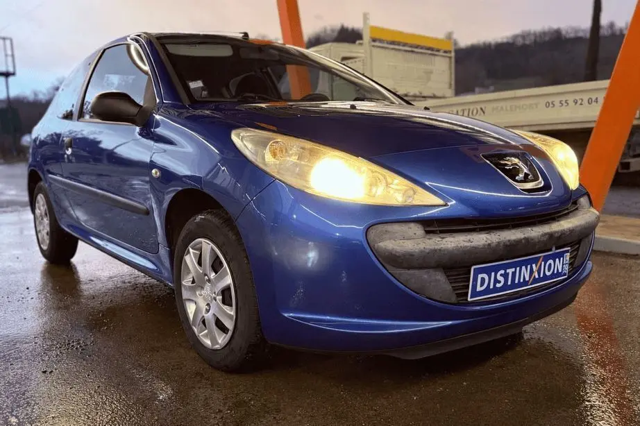 Peugeot 206+ bleu vue 3/4 avant droit avec phares allumés sur sol mouillé, devant structure orange.