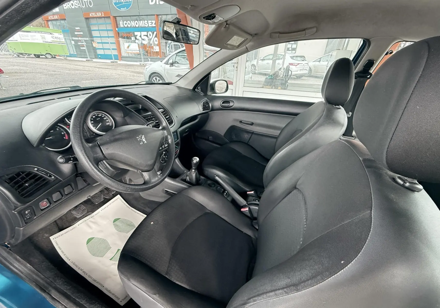 Vue intérieure côté conducteur d'une Peugeot 206+ bleue de 2011 avec sièges tissu noir et volant cuir.