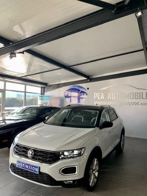 Volkswagen T-Roc blanc en 3/4 avant droit dans un showroom avec calandre noire et phares LED allumés.