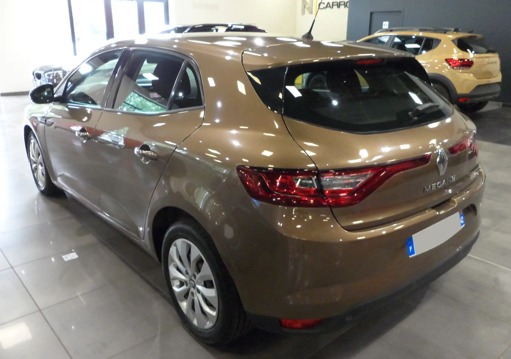 Vue 3/4 arrière gauche d'une Renault Mégane IV TCe 100 Life brun cappuccino en intérieur showroom.