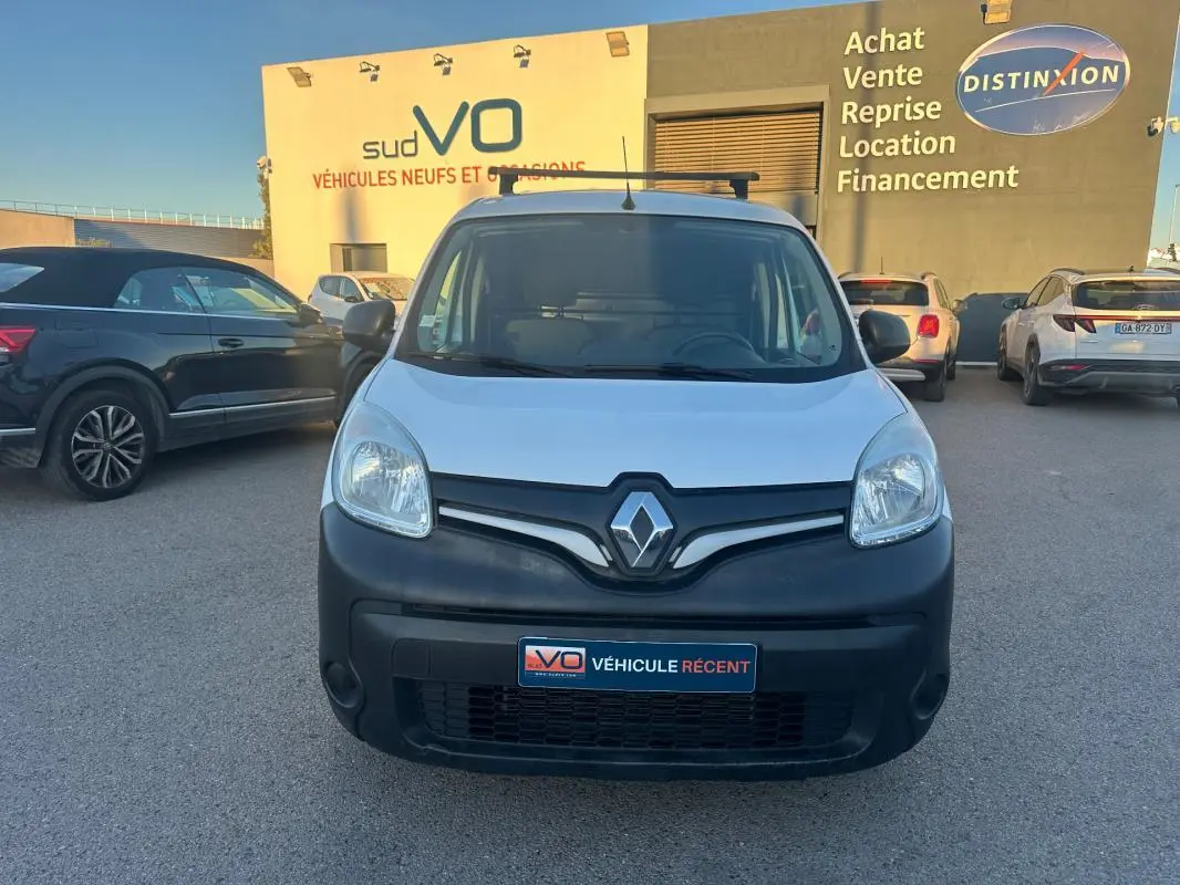 Vue frontale d'un Renault Kangoo Express blanc avec pare-chocs noir mat, stationné devant un bâtiment commercial.