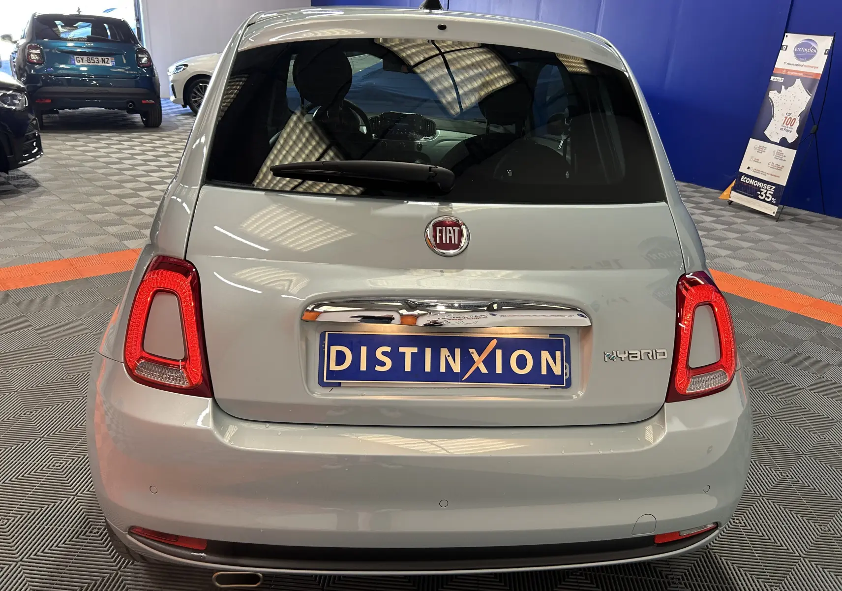 Vue arrière d'une FIAT 500 hybride couleur vert clair Rugiada, avec feux arrière allumés et plaque Distinxion visible en intérieur showroom.